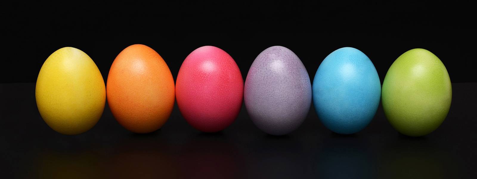 Six vibrant eggs in orange, pink, blue, green, purple, and yellow on a dark surface.