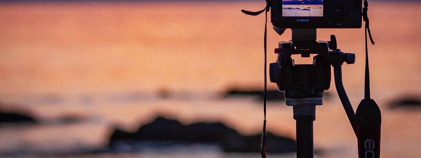 Camera on tripod capturing a sunset over the water with rocks in the foreground.
