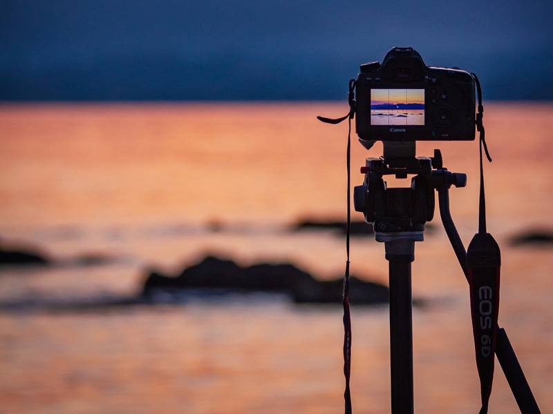 Camera on tripod capturing a sunset over the water with rocks in the foreground.