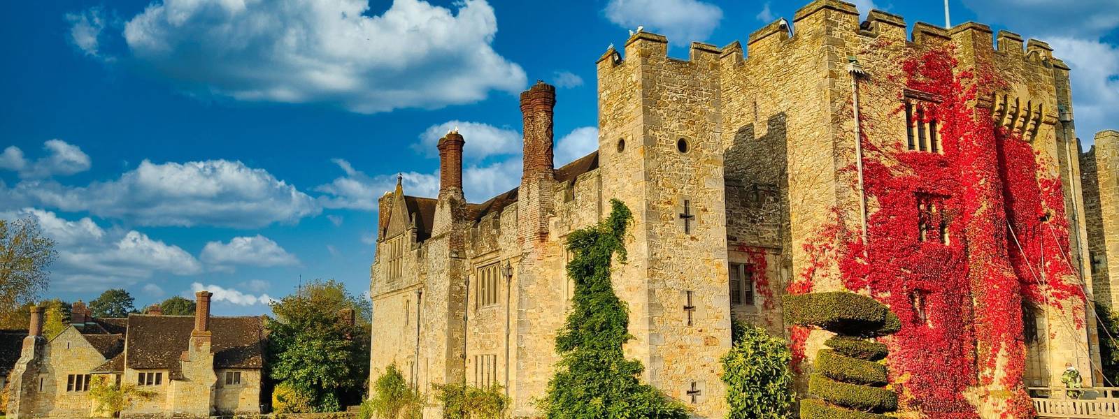 Historic Hever castle surrounded by greenery and a reflective pond under a blue sky.
