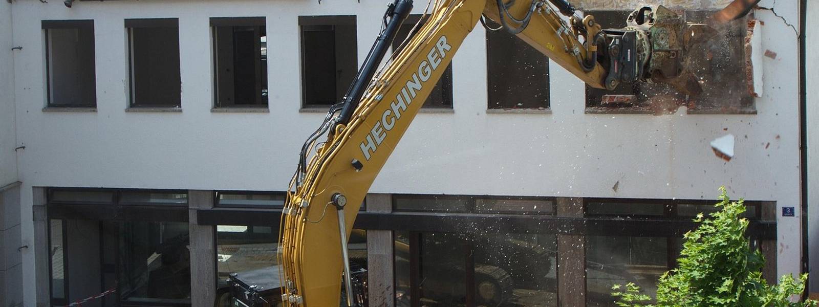 Excavator demolishing a building, debris falling from the roof in a construction site.