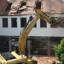 Excavator demolishing a building, debris falling from the roof in a construction site.